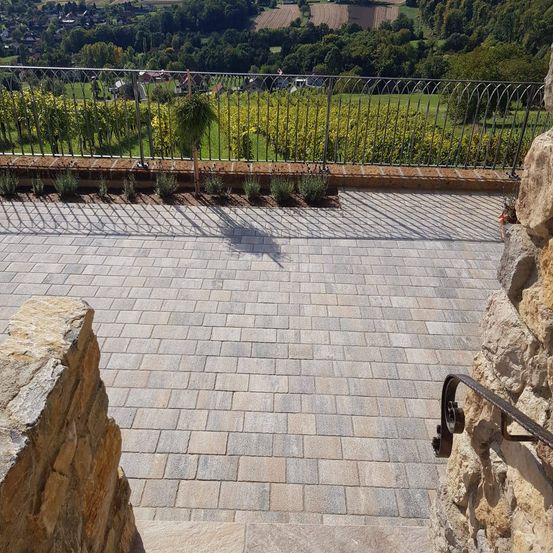 Eine gepflasterte Terrasse mit Metallgeländer bietet Blick auf Weinberge und die Landschaft. Schatten fallen auf den Weg.