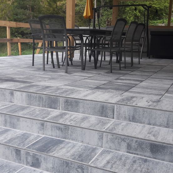 Eine Terrasse mit grauen Fliesen, einem Holzzaun und einem Tisch mit schwarzen Stühlen. Ein Regenschirm ist in der Nähe, und Bäume und Pflanzen sind im Hintergrund.