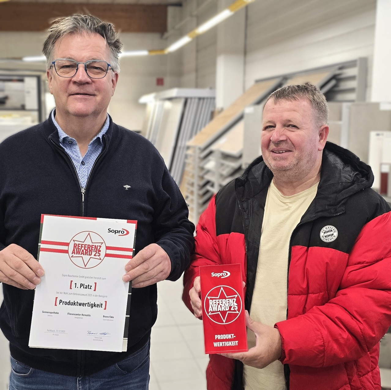 Zwei Männer stehen in einem Innenraum, beide halten Auszeichnungen in den Händen. Der Mann auf der linken Seite trägt einen blauen Pullover und eine Brille und hält ein Zertifikat mit den Worten 'Referend Award 25' und '1. Platz Produktwertigkeit'. Der Mann auf der rechten Seite trägt eine rote Jacke und hält eine rote Box mit den gleichen Worten. Sie lächeln beide.