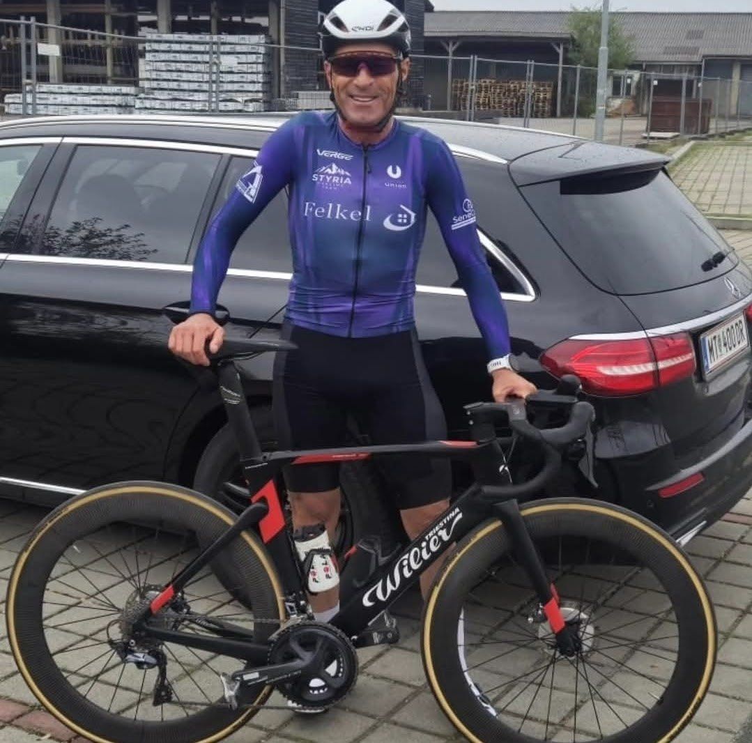 Ein Mann in Radsportbekleidung steht neben seinem Fahrrad, ein schwarzes Auto dahinter.