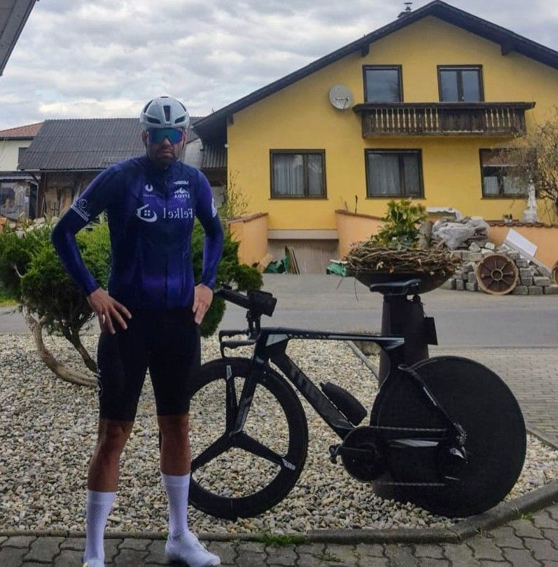 Ein Mann in Radbekleidung, mit Helm und Sonnenbrille, steht neben einem schwarzen Fahrrad auf einem Kiesweg vor einem gelben Haus.