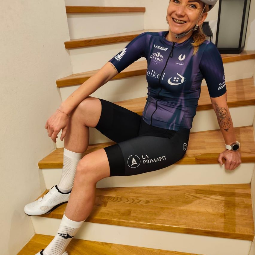 Eine Frau in einem Radtrikot mit dem Wort 'Verge' und dem Markenlogo von 'Styrian' sitzt auf den Holzstufen, lächelt und posiert für ein Foto.