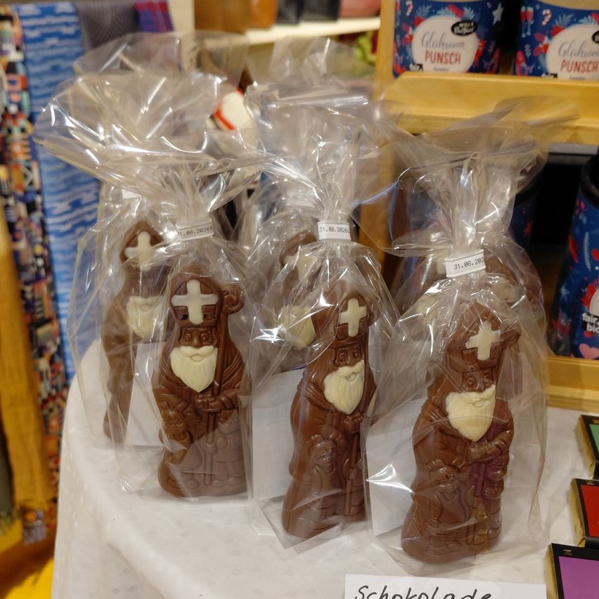 Ein Tisch zeigt mehrere Schokoladenfiguren von Sankt Nikolaus, jede in Plastik verpackt, mit Preisschildern und einem Schild Schokolade. Dahinter befinden sich Regale mit verschiedenen Artikeln.