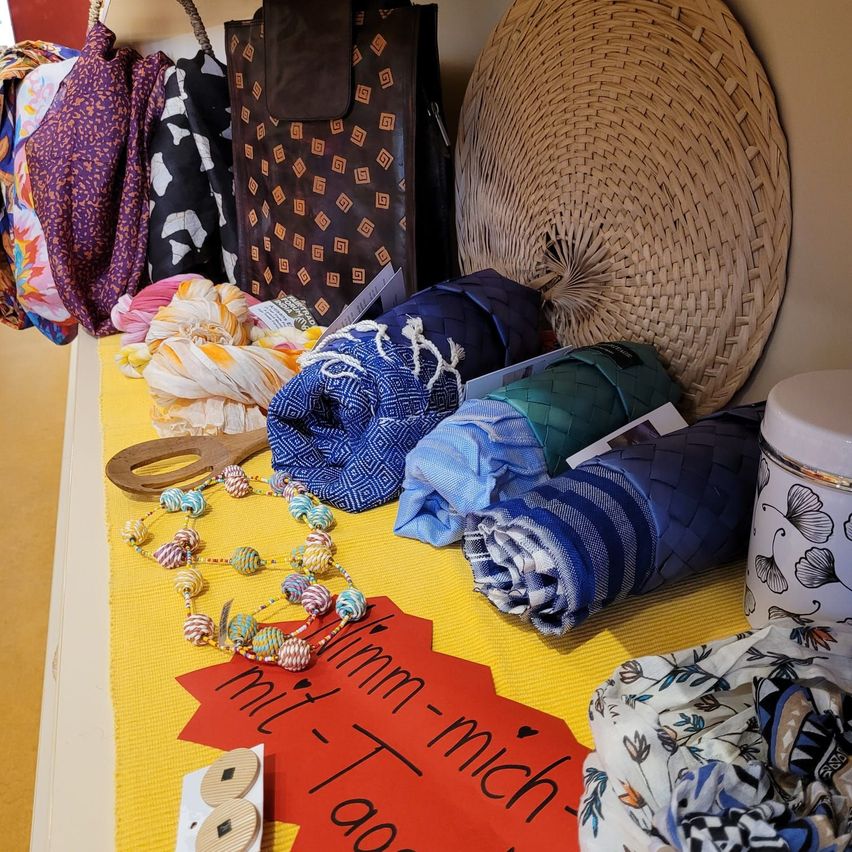 Ein gelber Tisch zeigt verschiedene Artikel wie Schals, einen Korb und eine Halskette. Ein rotes Papier mit den Worten 'mit ihm mich' ist an den Tisch geheftet.