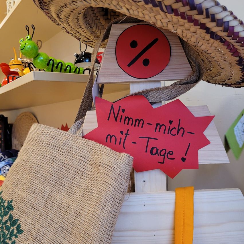 Ein Strohhut und eine gewebte Tasche hängen an einem Holzschild mit Rabattsymbol. Dahinter steht ein Regal mit bunten Gegenständen.