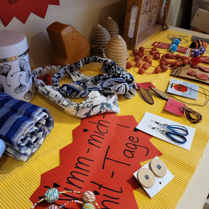 Ein Tisch zeigt eine Vielzahl von handgefertigten Artikeln, darunter Schals, Perlenketten und Ohrringe. Ein Glas mit Blumendesign steht links, und ein herzförmiges Schild mit der Aufschrift 'umm-mich-Tage'. Der Tisch ist mit einem gelben Tischtuch bedeckt.