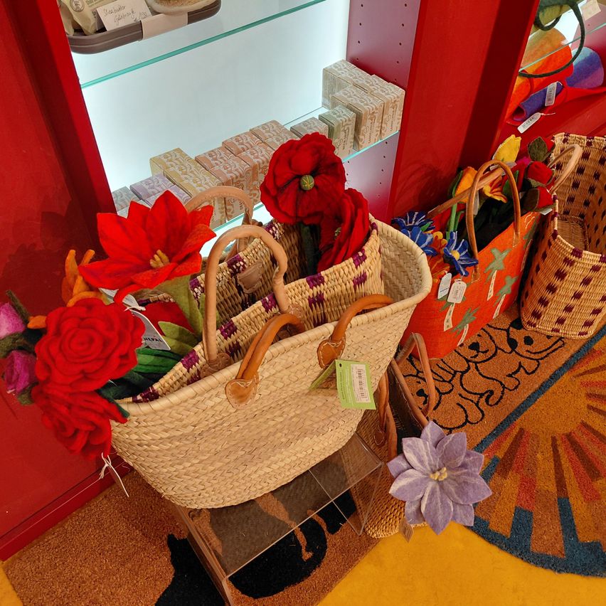 A basket filled with red and orange flowers sits on a glass stand. Nearby, two other baskets, one orange with colorful designs and the other woven, are on a carpet. A red shelf with products is visible in the background.
