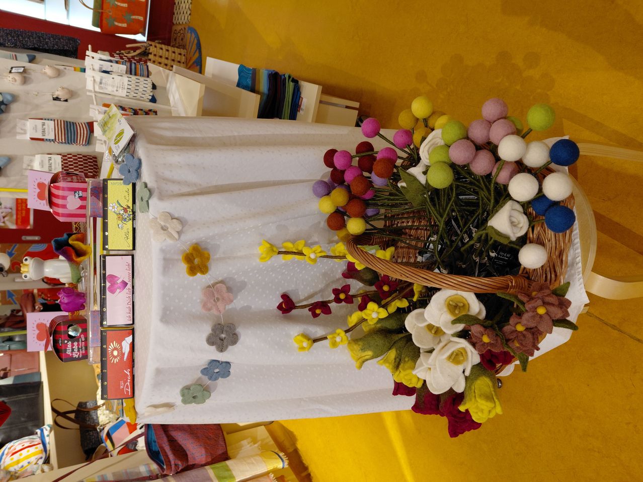A table with a white tablecloth has a woven basket filled with colorful felt flowers. Various items are displayed on shelves behind the table.