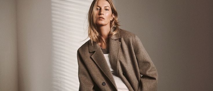 Eine Frau mit blonden Haaren steht vor einem Fenster, trägt einen braunen Mantel und ein weißes Shirt und schaut zur Seite.