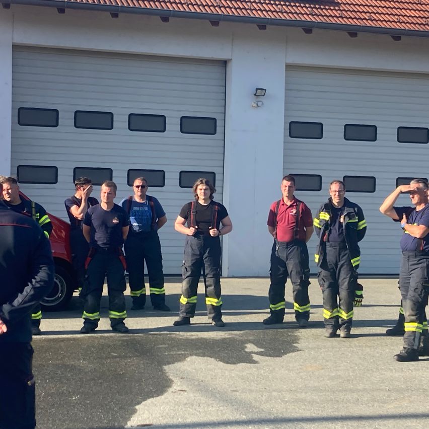 Eine Gruppe von Feuerwehrleuten steht vor einer Feuerwache mit einem roten Auto in der Nähe. Die Feuerwehrleute tragen Uniformen und einige haben ihre Hände auf der Stirn zum Gruß.