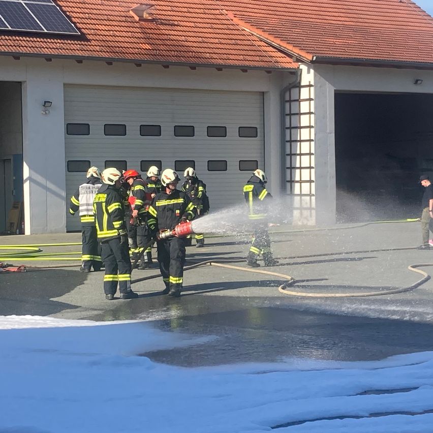 Feuerwehrleute in Uniform stehen in der Nähe einer Feuerwache und sprühen mit einem Schlauch Wasser. Ein Mann in einem schwarzen T-Shirt steht hinter ihnen.