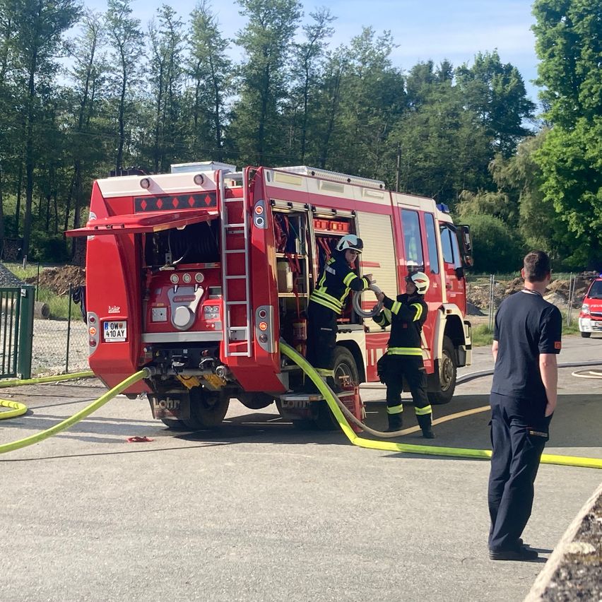 Zwei Feuerwehrleute verbinden einen Schlauch mit einem roten Feuerwehrauto. Ein Mann in einem schwarzen Shirt steht in der Nähe.