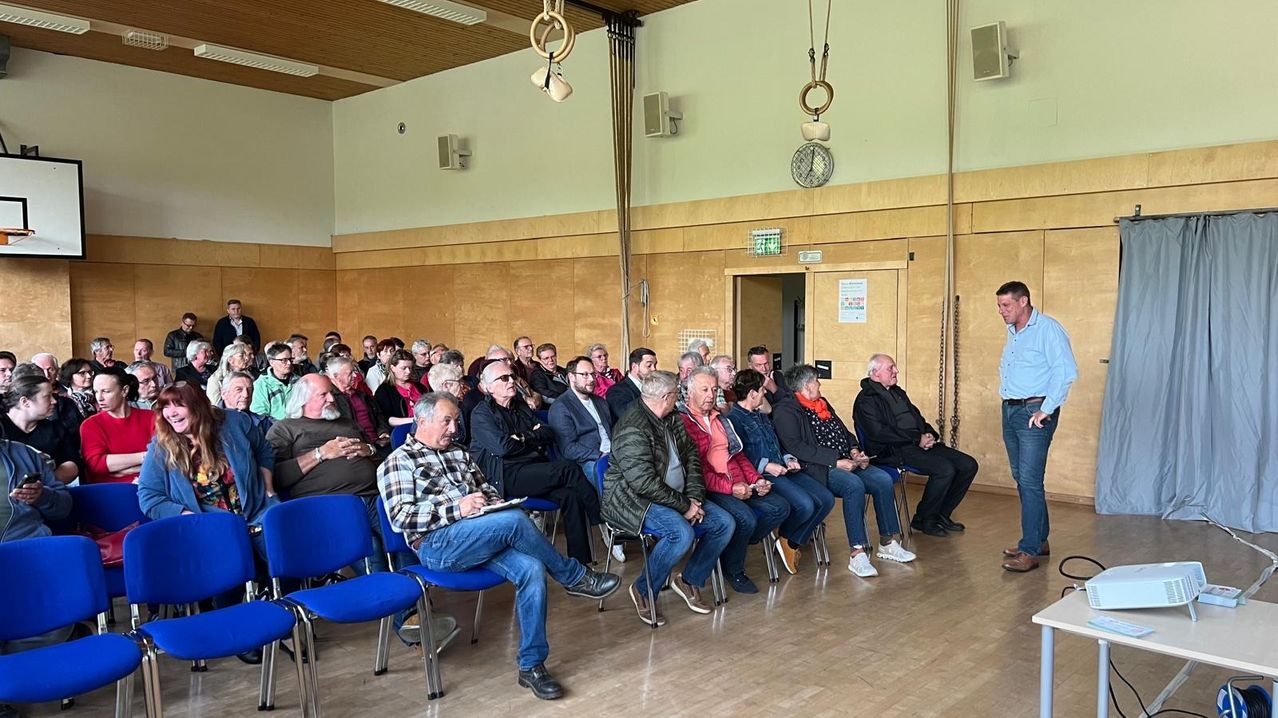 Eine Gruppe von Menschen, hauptsächlich ältere Menschen, sitzen in einer Reihe von Stühlen in einem Saal und lauschen einem Sprecher. Der Raum hat Holzböden und Wände.