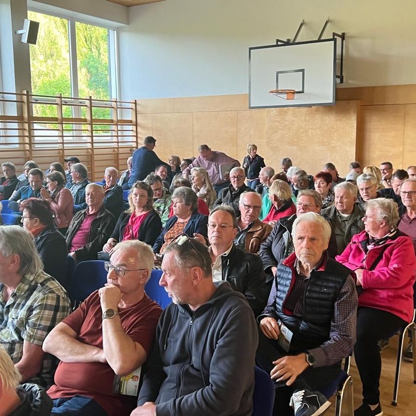 Eine große Menschenmenge sitzt in Stühlen in einer Turnhalle. Sie beobachten aufmerksam eine Präsentation. Der Raum hat Holzwände und große Fenster, durch die helles Sonnenlicht scheint.