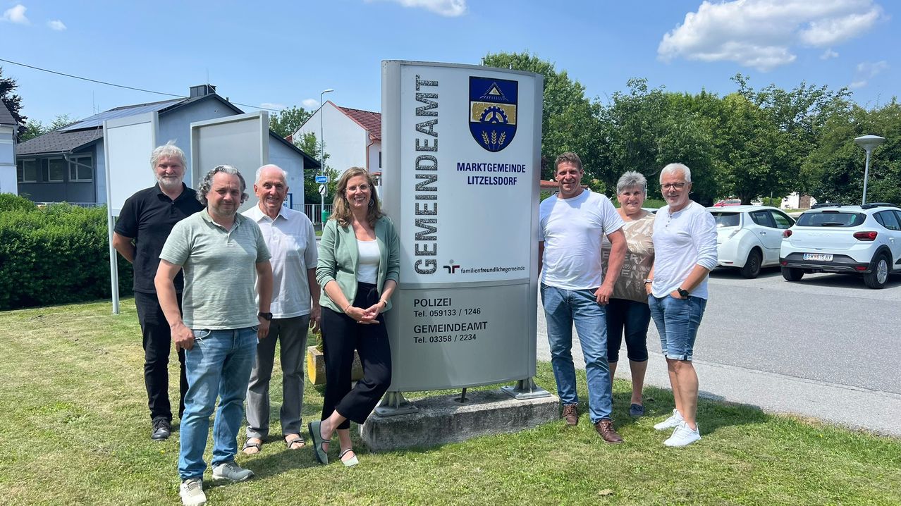 Eine Gruppe von Menschen, einschließlich einer Frau, steht vor einem Gemeindeschild in Litzelsdorf. Sie lächeln für ein Foto.