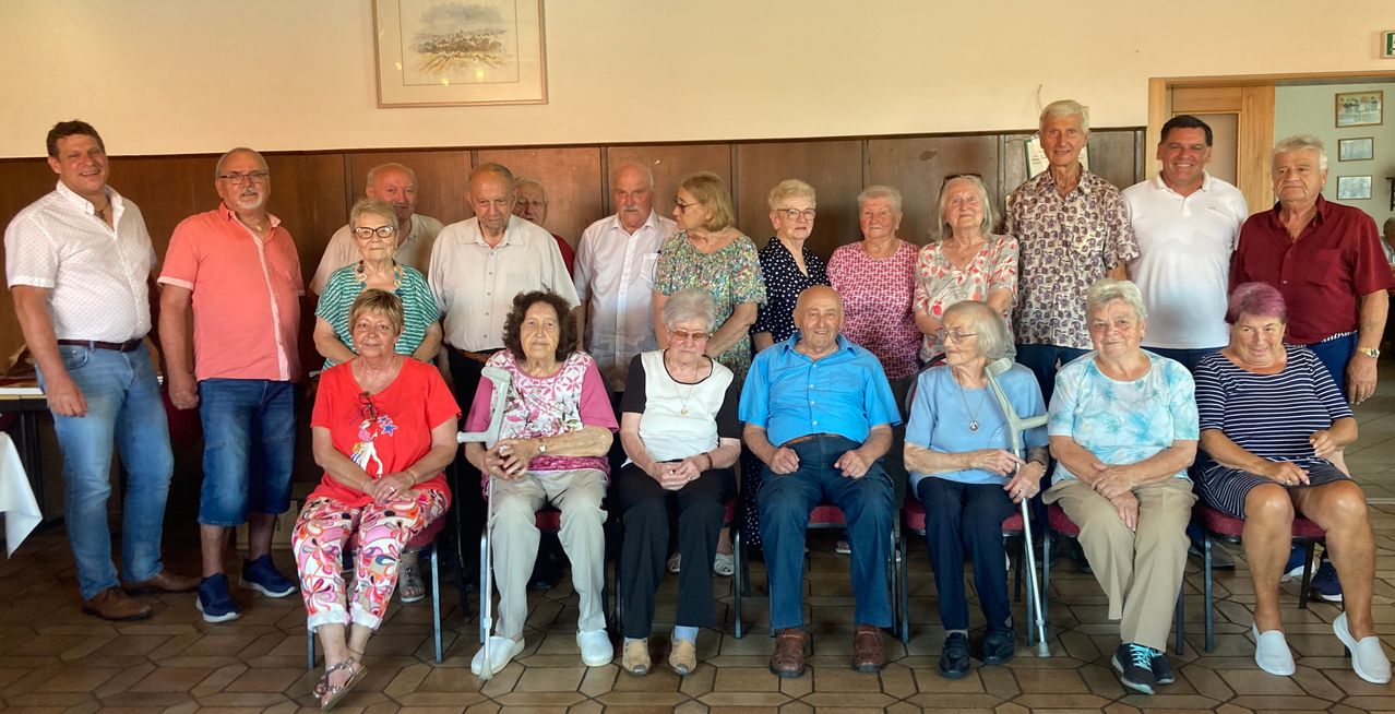 Eine Gruppe älterer Menschen posiert für ein Foto. Sie sitzen und stehen in einem Raum mit gefliesten Böden und Holzwänden.