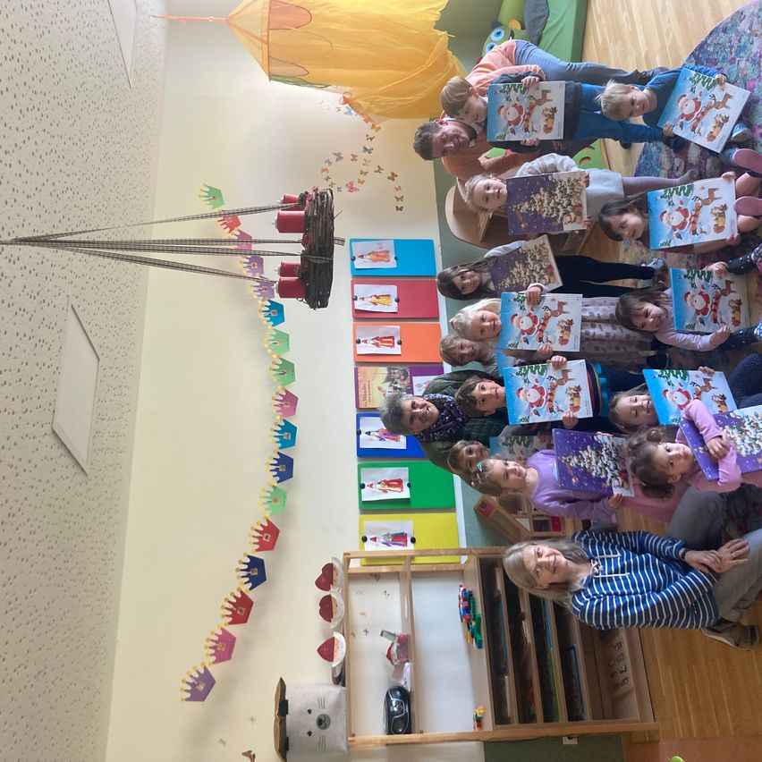 Eine Gruppe von Kindern und Erwachsenen posiert für ein Foto in einem Raum mit Weihnachtsdekoration und einer Laterne. Die Kinder halten Bücher und die Erwachsenen lächeln.