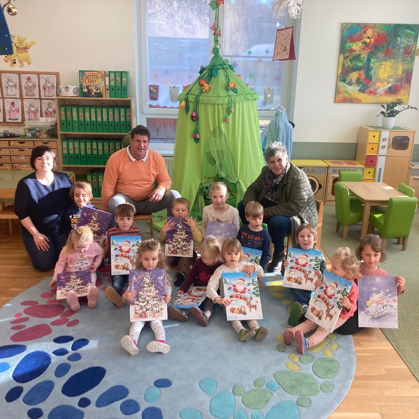 Eine Gruppe von Kindern und Erwachsenen in einem Raum, lächelnd und Weihnachtskarten haltend. Dahinter ein grünes Zelt und ein Gemälde an der Wand. Regale mit Büchern und ein Tisch mit Stühlen.