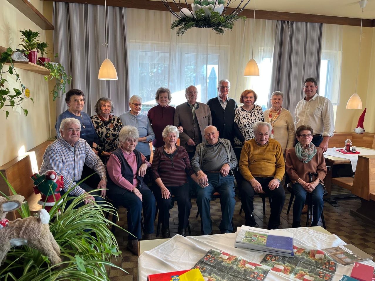 Eine Gruppe älterer Personen, gekleidet in verschiedene Winterkleidung, versammelt sich in einem Raum. Sie sitzen um einen Tisch, der mit einem weißen Tuch und Büchern bedeckt ist. Eine festliche Dekoration hängt über ihnen.