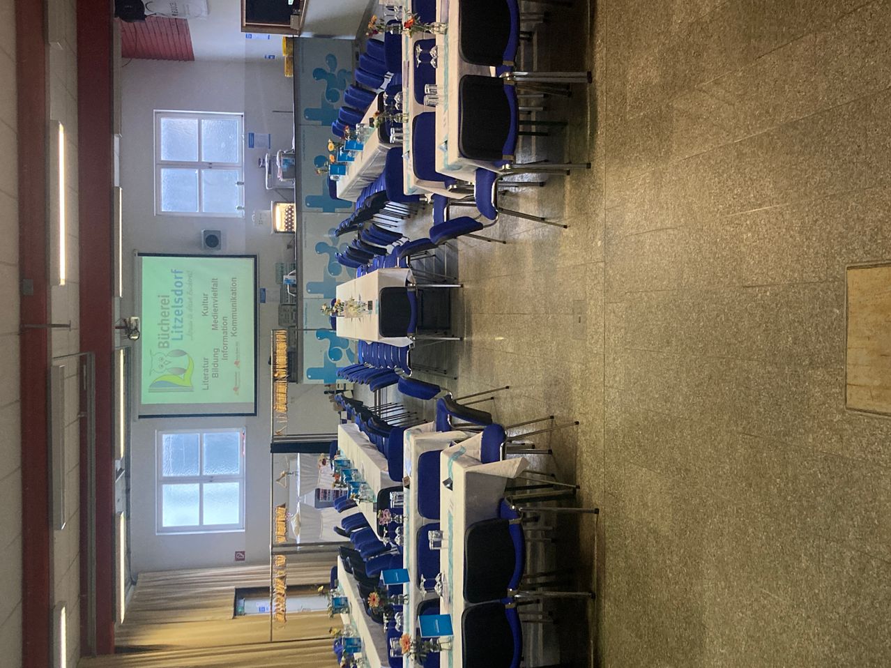 Eine Vogelperspektive eines Raumes mit Reihen von Tischen und Stühlen für eine Versammlung. Die Tische haben weiße Tischdecken und blaue Blumen. Eine Projektionsleinwand ist links mit einer Präsentation.