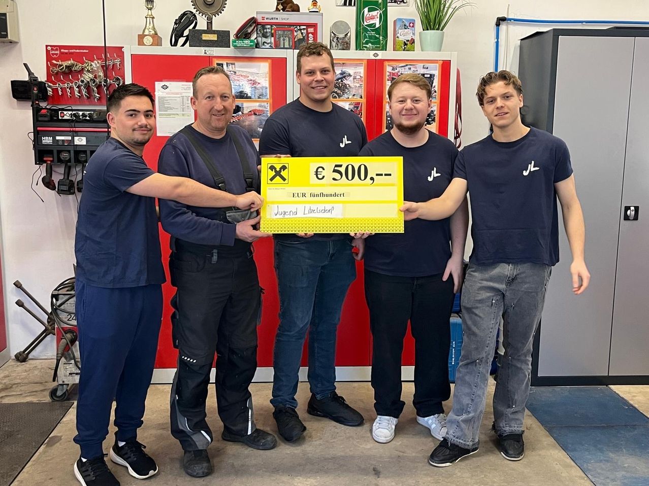 Fünf Männer in blauen Uniformen stehen in einem Raum und halten einen gelben Scheck über 500 Euro. Sie lächeln und scheinen zu feiern.