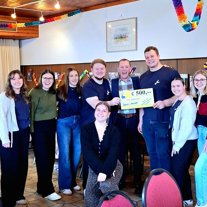 Eine Gruppe von Menschen posiert für ein Foto in einem Raum. Eine Frau in der Mitte hält ein Zertifikat für 500 Euro. Hinter ihr lächeln zwei Männer. Der Raum hat Holzwände und eine Decke mit Lichtern.