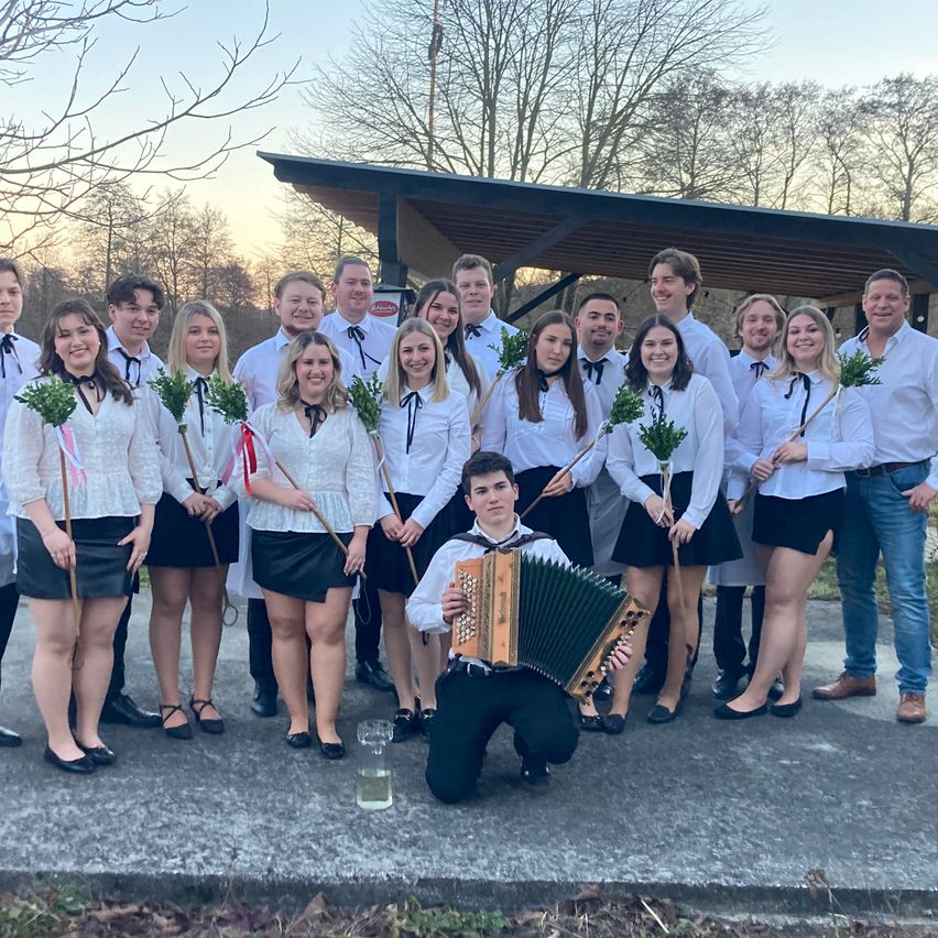 Eine Gruppe von Musikern und Tänzern posiert für ein Foto im Freien. Ein junger Mann kniet mit einem Akkordeon. Die anderen halten Stöcke mit grünen Blättern. Sie tragen traditionelle Kleidung.