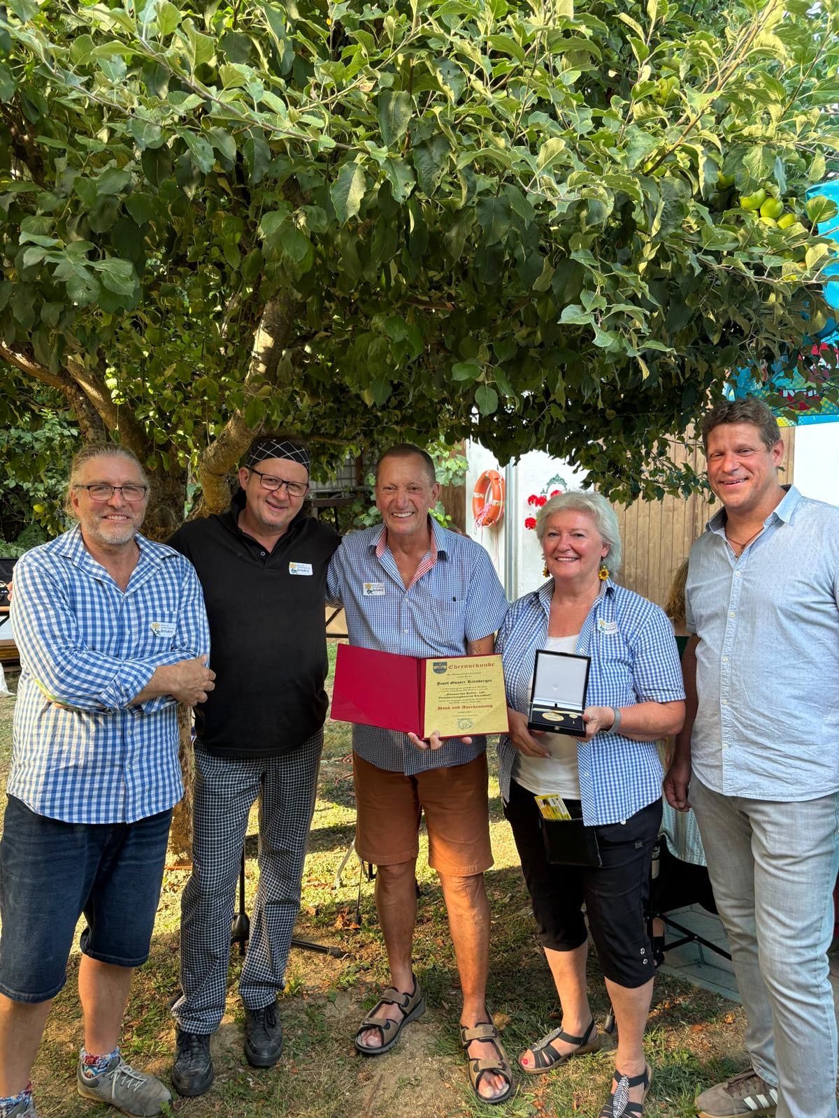 Five adults stand under a tree, smiling for a photo. The man in the center holds a red folder and a medal. The woman next to him holds a medal. The man on the right wears a necklace. The man on the left wears glasses.