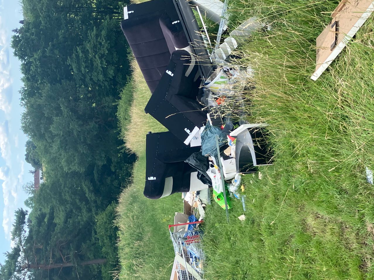 Eine Luftaufnahme zeigt einen großen Haufen Möbel und Müll in einem Grasfeld, umgeben von Bäumen.