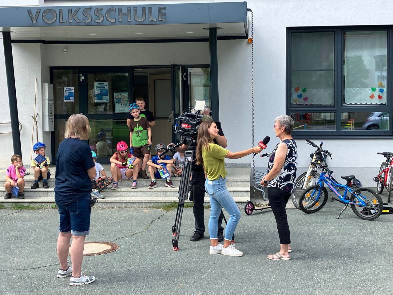 Ein junges Mädchen spricht in ein Mikrofon, das von einer erwachsenen Frau gehalten wird. Sie stehen vor einer Schule, wo Kinder auf den Stufen sitzen und Fahrräder geparkt sind.