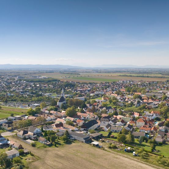 Eine Luftaufnahme einer Stadt mit zahlreichen Häusern, Bäumen und einer Kirche in der Mitte. Die Landschaft zeigt weite grüne Felder und entfernte Berge unter einem klaren blauen Himmel.