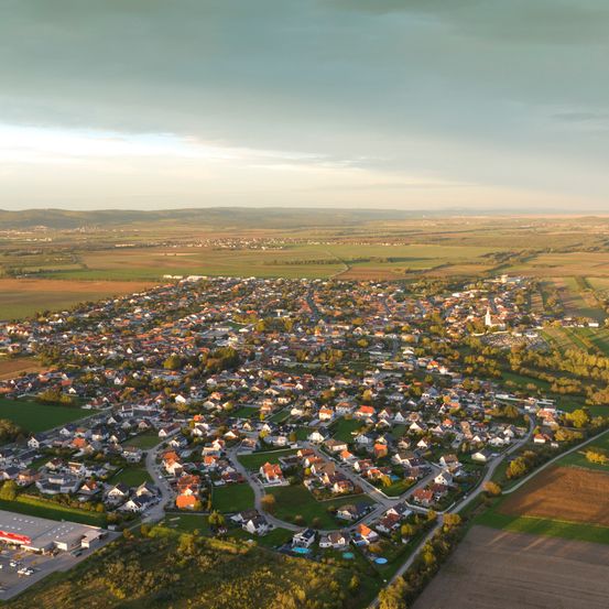 Luftaufnahme einer Vorstadt mit vielen Häusern, grünen Feldern und einigen Bäumen. Der Himmel ist bewölkt.