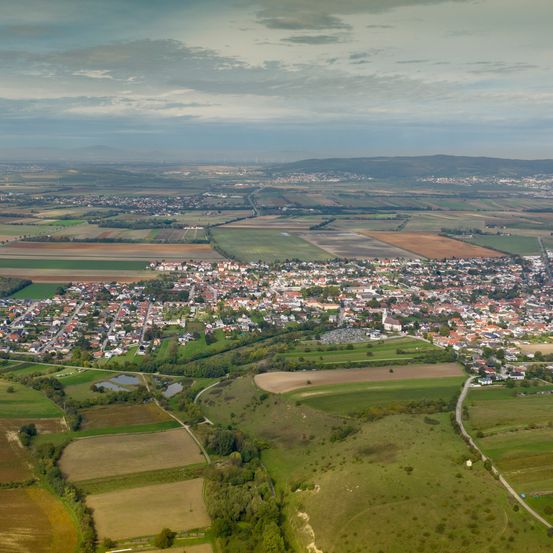 Luftaufnahme einer Stadt umgeben von grünen Feldern und kleinen Hügeln. Die Stadt hat viele Gebäude, einige mit roten Dächern. Die Felder befinden sich in verschiedenen Wachstumsphasen, und einige haben Wassertümpel.