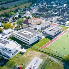 Luftaufnahme eines Vorstadtviertels mit mehreren modernen Gebäuden, einem Spielplatz und einem Fußballfeld. Die Gebäude haben Solarmodule auf ihren Dächern.