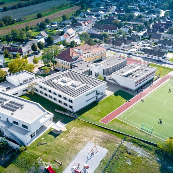 Luftaufnahme eines Vorstadtviertels mit mehreren modernen Gebäuden, einem Spielplatz und einem Fußballfeld. Die Gebäude haben Solarmodule auf ihren Dächern.