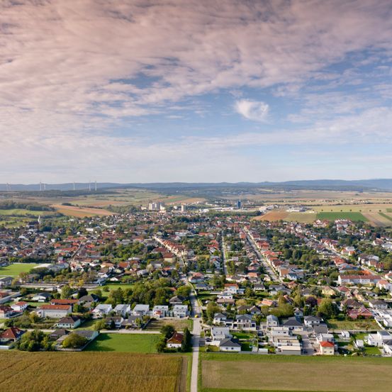 Eine Luftaufnahme einer Stadt mit vielen Häusern und Gebäuden. Es gibt auch Bäume und Felder. Der Himmel ist bewölkt. In der Ferne befinden sich Windräder.