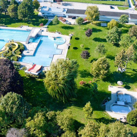 Luftaufnahme eines großen Außenpoolbereichs mit einer Rutsche, Sprungbrettern und einer umliegenden Rasenfläche. Bäume und Sträucher sind verstreut. Ein Gebäude mit Dach ist im Hintergrund.