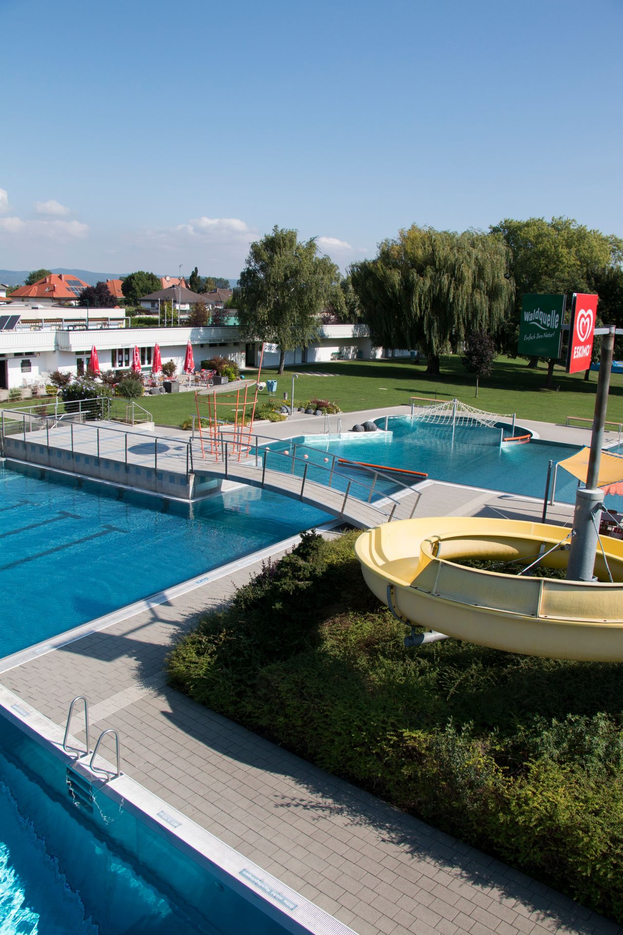 Ein Luftbild eines großen Schwimmbadkomplexes mit einer Brücke, Wasserrutsche und einem roten Schild mit 'Waldquelle' darauf.