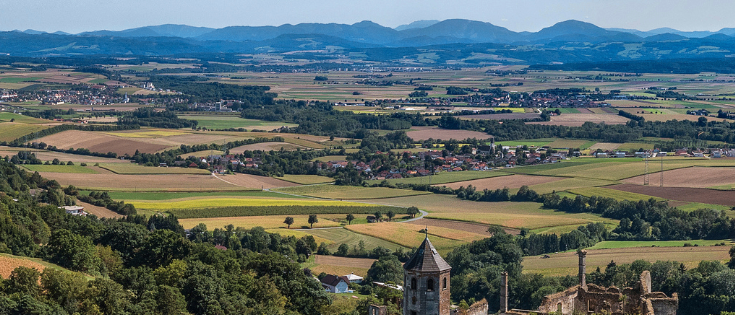 Luftaufnahme einer ländlichen Landschaft mit Feldern, Bergen in der Ferne und einem Dorf in der Mitte. Die Landschaft ist mit Bäumen und Grünflächen übersät.