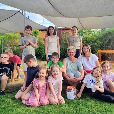 Eine Gruppe von Kindern und Erwachsenen versammelt sich auf dem Rasen unter einem Baldachin. Einige sitzen, andere stehen, und ein Junge bläst Seifenblasen. Ein Glas ist auf dem Rasen platziert.