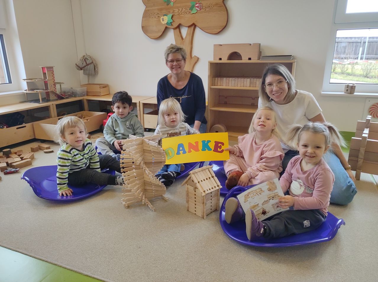 In einem Klassenzimmer sitzen mehrere Kinder und zwei Frauen auf blauen Schlitten. Ein Kind hält ein Schild mit der Aufschrift Danke. Dahinter steht ein Holzbaum und Regale mit Spielzeug.