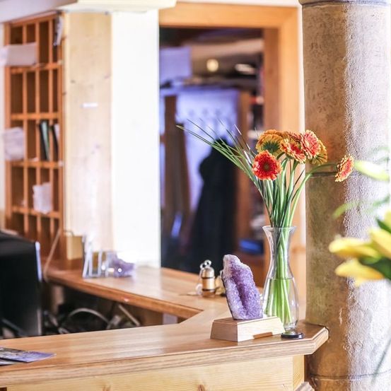 A wooden counter with flowers and a purple rock. A shelf with books and papers. A blurry figure in a room.