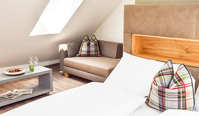 Ein gemütliches Schlafzimmer mit einem Bett, einer Couch und einem Couchtisch. Das Bett hat karierte Kissen und die Couch einen karierten Überwurf. Der Raum hat einen hölzernen Kopfteil, ein Fenster und einen kleinen Teller mit Essen auf dem Tisch.