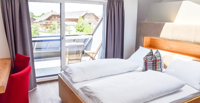 Ein Schlafzimmer mit einem großen Bett, weißen Kissen und einem Balkonblick auf ein Haus und eine Terrasse mit Stühlen.