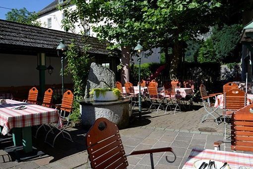 Ein Freiluftrestaurant mit Holztischen, Stühlen und Bäumen. In der Mitte steht ein Steinpfeiler mit einer Topfpflanze. Die Tische sind mit rot-weiß karierten Tischdecken bedeckt.