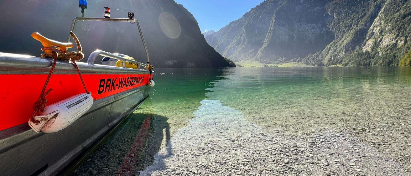 Ein Boot mit der Aufschrift BRK-Wasserwacht liegt am Ufer eines Sees mit klarem Wasser. Im Hintergrund befinden sich Berge.
