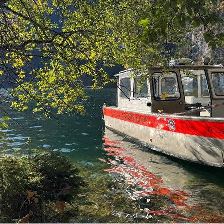 Ein Boot mit rotem Streifen ist in einem See verankert. Es ist von Bäumen mit grünen Blättern umgeben. Das Wasser ist ruhig, und die Reflexion des Bootes ist im Wasser sichtbar.