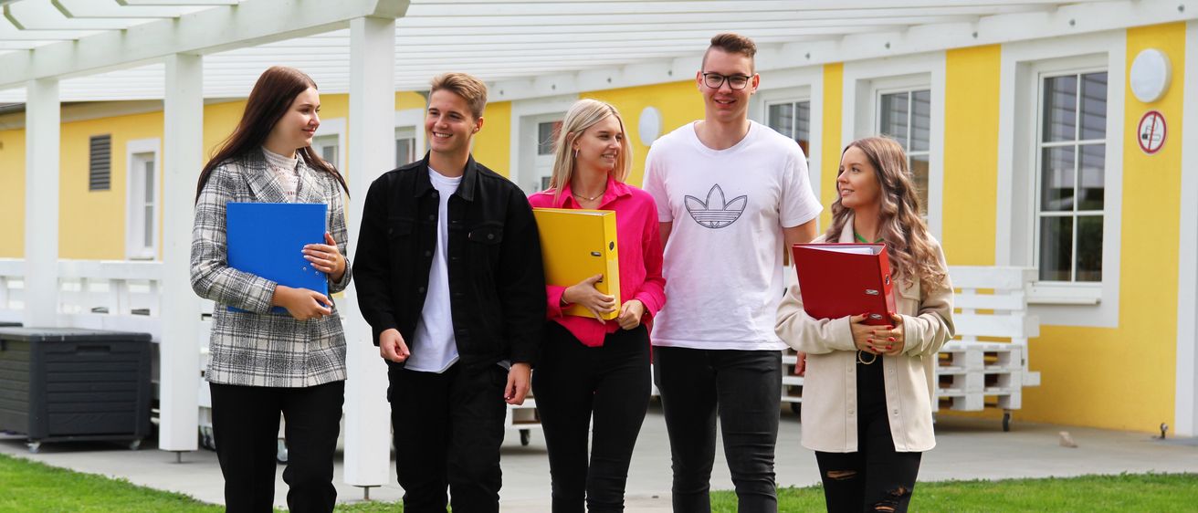Fünf junge Erwachsene in lässiger Kleidung stehen auf einem Bürgersteig und halten verschiedene farbige Ordner in den Händen. Sie scheinen Schüler zu sein, wobei einer eine Brille trägt und eine andere einen blauen Ordner hält. Sie lächeln und posieren für ein Foto vor einem Gebäude mit gelben Wänden und weißen Einfassungen.