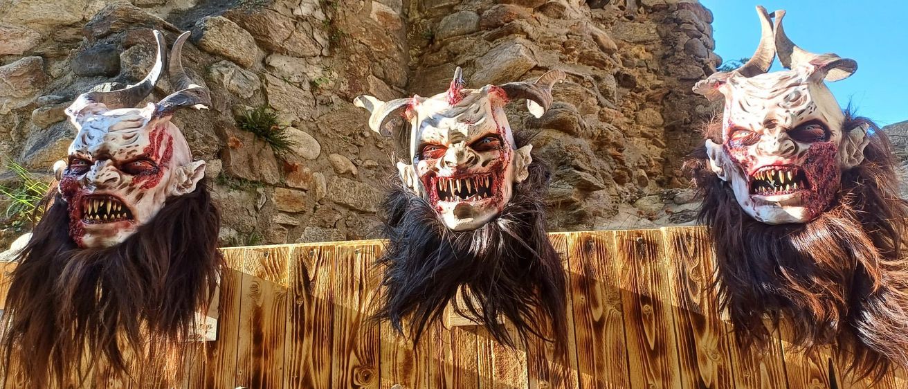 Drei Masken mit Hörnern und Blutspuren hängen an einem Holzzaun vor einer Steinmauer, mit einem Blick auf einen Baum durch ein Loch.
