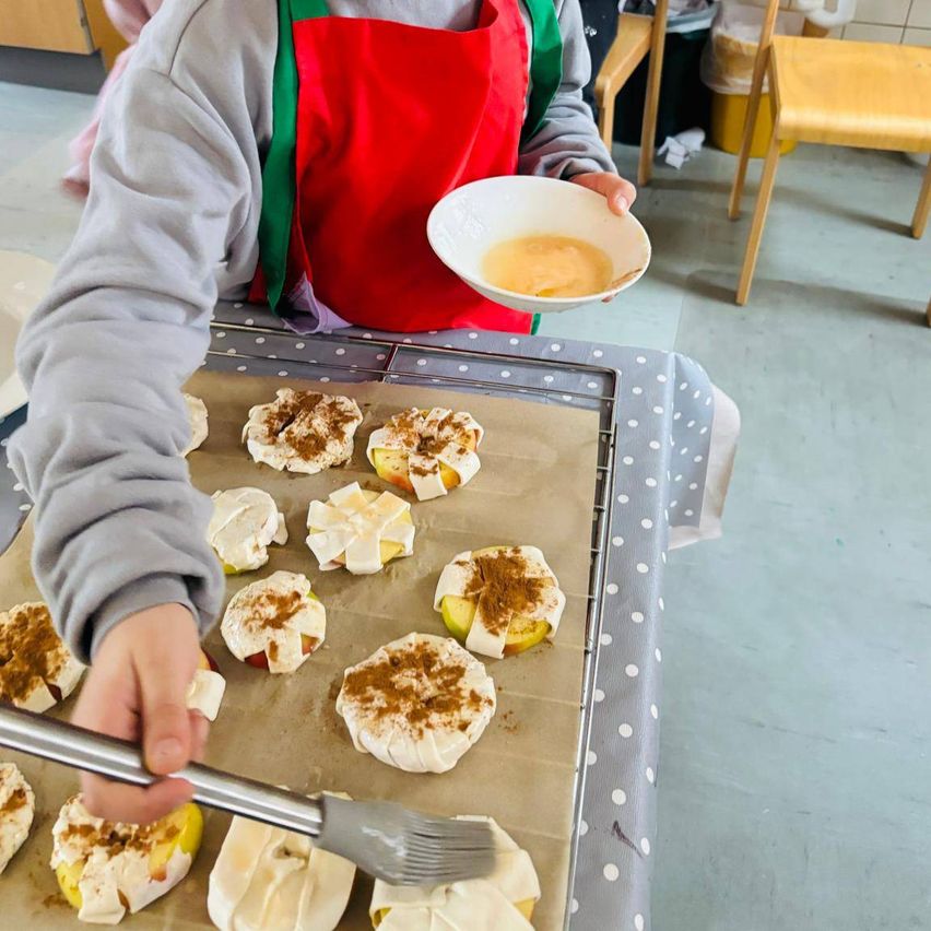 A child is holding a bowl of mixture, probably in a kitchen. The child is wearing a gray sweatshirt and a red apron. The child is holding a brush and seems to be brushing the mixture on the food. The food is placed on a tray with a polka-dot cover.